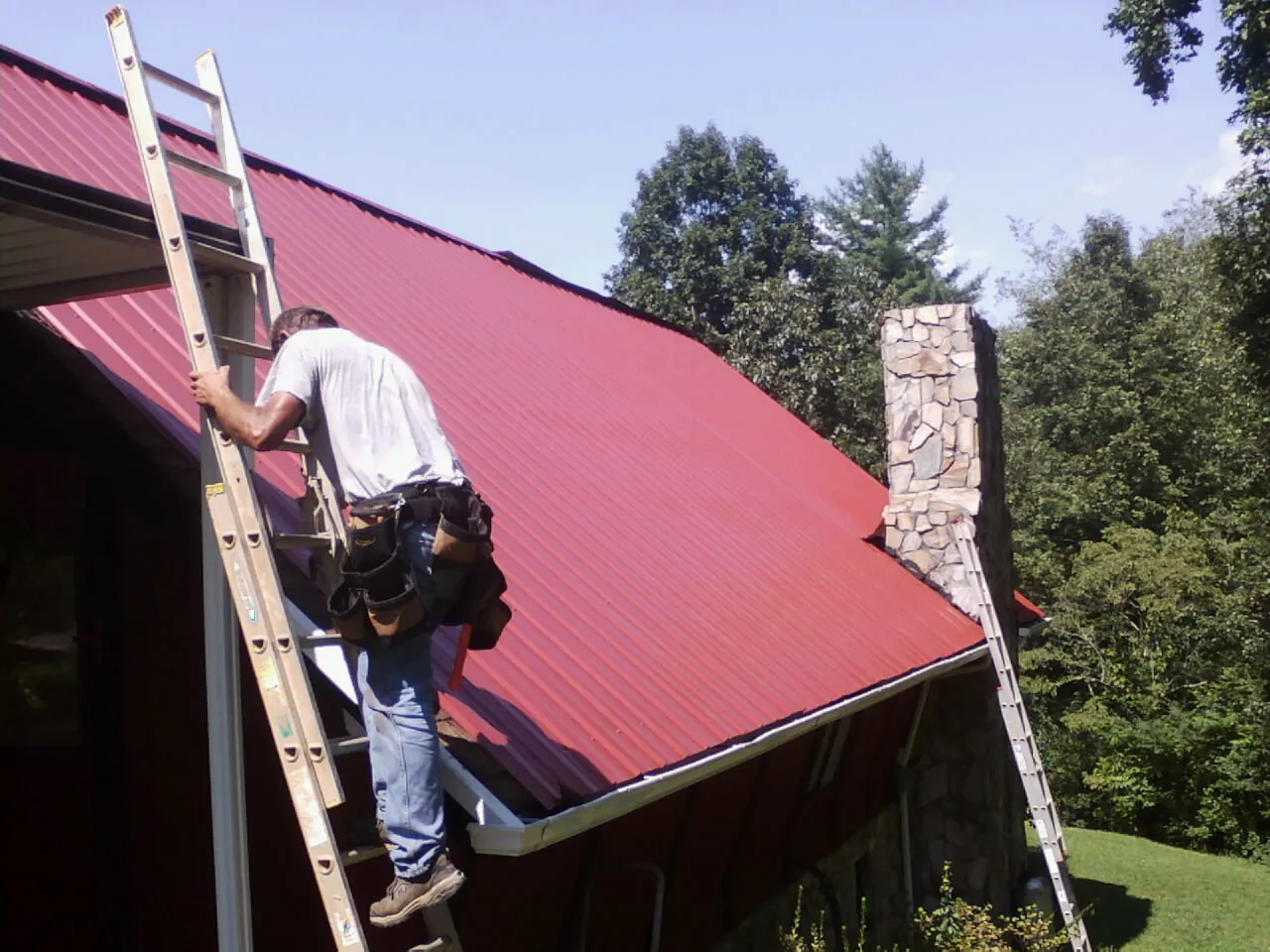 Roofer on a ladder beside a red metal roof and stone chimney — Soffit & Fascia Repair by Roofing Brothers in Abita Springs